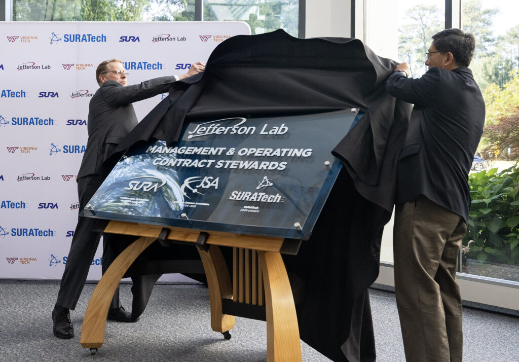 Two men unveiling a large blue plaque that reads "Jefferson Lab Management & Operating Contract Stewards" with the following logos and dates: SURA (1984-2006), Jefferson Science Associates (2006-2026), SURATech (2026-present).
