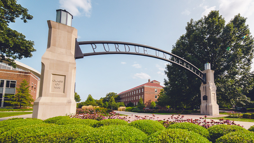 A metal arch that reads "Purdue University" held by two stone pillars in front of Purdue campus buildings and green trees. The image includes a blue sky and green bushes in front of the arch.