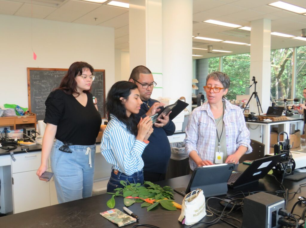 Three young interns stand standing notes and listening to a woman. All stand at a lab bench with computers, equipment, and leaf samples.